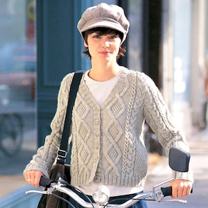 May include: A light grey cable-knit cardigan with a V-neck and button closure. The cardigan features a diamond pattern and is worn with a grey newsboy cap and a white t-shirt. A black shoulder bag is also visible.