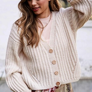 Könnte beinhalten: Cremefarbene, grob gestrickte Strickjacke mit V-Ausschnitt und Holzknöpfen. Der Pullover hat eine gerippte Textur und lange Ärmel. Eine goldene Halskette ist sichtbar. Die Strickjacke ist Teil eines lässigen Outfits.