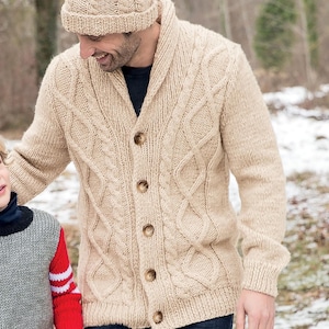 May include: A beige cable knit cardigan with a shawl collar and button closure. The cardigan is worn over a white t-shirt and blue jeans. The person is wearing a beige knit beanie with a brown pom-pom.