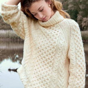 May include: Cream-colored, cable-knit turtleneck sweater. The sweater features a textured pattern with diamond and cable designs. The thick knitwear has a high, ribbed collar and long sleeves. The overall style is cozy and warm.
