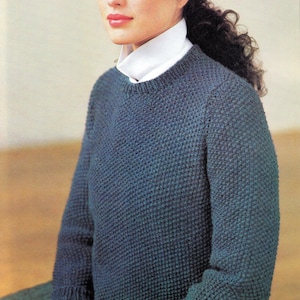 Könnte beinhalten: Ein dunkelblaugrüner Strickpullover mit strukturiertem Muster. Der Pullover hat lange Ärmel und einen Rundhalsausschnitt, getragen über einem weißen Hemd mit Kragen. Ein schwarzer Hut wird auf dem Kopf getragen.