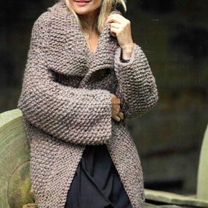 Könnte beinhalten: Ein langer, offener Strickmantel in einem neutralen Taupe-Ton. Der Mantel hat einen breiten, schalkragenartigen Kragen und lange Ärmel. Die grob gestrickte Textur verleiht diesem gemütlichen Oberbekleidungsstück einen optischen Reiz.