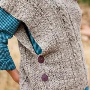 May include: A knitted vest in a neutral color with a textured pattern and cable knit detailing. The vest has three large, dark buttons down the side. A blue plaid shirt is visible underneath.