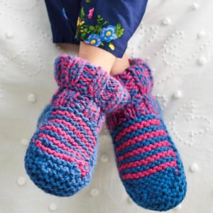 May include: A pair of cozy, hand-knitted slippers in blue and pink. The slippers feature a striped pattern in pink and blue, with a folded-over cuff. The image shows the slippers on a white surface, with a glimpse of blue pants with floral embroidery.