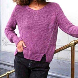 May include: A purple V-neck sweater with long sleeves. The sweater has a ribbed texture and a split hem. The person is wearing black jeans. The sweater is a solid color.