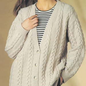 Può includere: Un cardigan beige chiaro a maglia a trecce con scollo a V e chiusura con bottoni. Il maglione a maniche lunghe presenta un design a trecce strutturato ed è indossato sopra una maglietta a righe bianche e nere. Il cardigan è di colore neutro.
