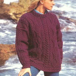 May include: A dark purple cable-knit sweater with a high neckline. The sweater features intricate cable patterns on the front and sleeves. The sleeves are long and the sweater is a loose fit. The person is wearing blue jeans.