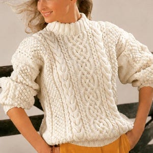 May include: An off-white, long-sleeved, chunky knit sweater featuring a high mock neck collar. The garment displays a complex texture with vertical cable knit patterns running down the center and sides, complemented by intricate bobble stitch details.