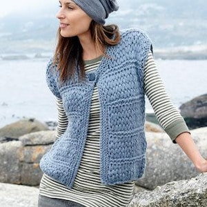Op de afbeelding: Een lichtblauw gebreid vest met een knoopsluiting. Het vest wordt gedragen over een gestreept shirt met lange mouwen en een groen shirt. De persoon draagt een grijze beanie. De achtergrond toont een water en rotsen.
