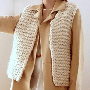 Peut inclure: Une écharpe en tricot épais de couleur crème drapée sur un blazer beige. L'écharpe est faite de fil épais, créant un aspect texturé et douillet. Le blazer a un design classique avec des revers et une fermeture à bouton. L'esthétique générale est minimaliste et élégante.