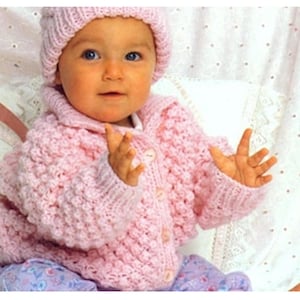 May include: A light pink knitted cardigan with a textured bobble stitch pattern, a wide collar, and front buttons, paired with a matching knitted beanie hat. The garment features long sleeves with ribbed cuffs and a soft, chunky knit construction.