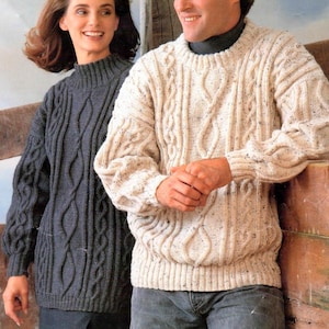 May include: Two cable-knit jumpers. One is a dark grey with a high neck, the other is off-white with a crew neck. Both jumpers have a textured, woven design. The jumpers are worn by two people.