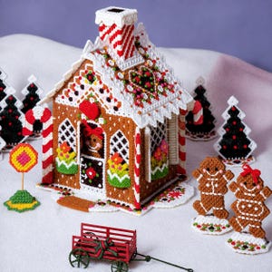 May include: A plastic canvas gingerbread house with white icing trim, a red and white striped chimney, and candy-like decorations. It is accompanied by two gingerbread figures, a small red wagon, and several black and white Christmas trees.