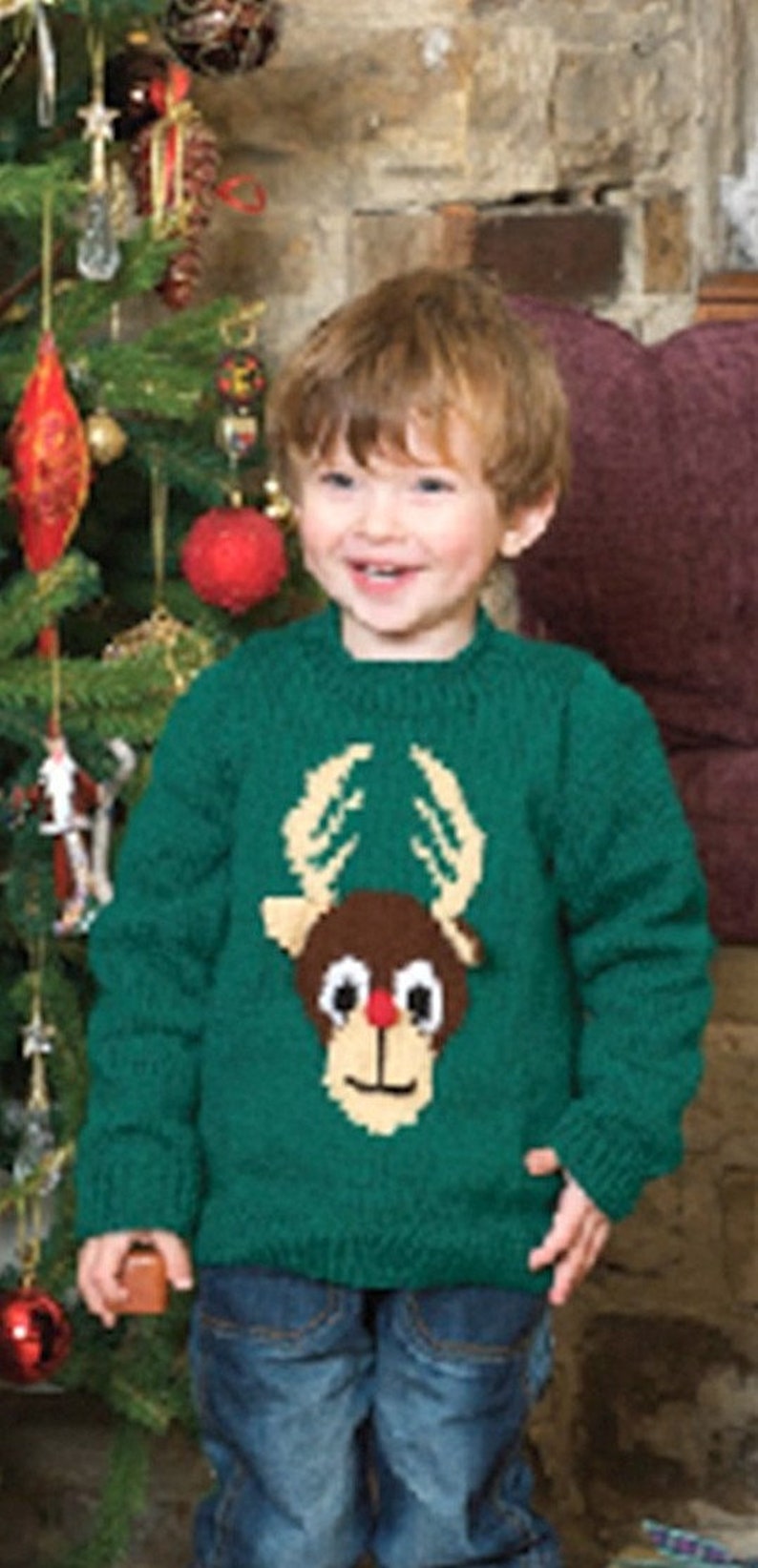 May include: A young child wearing a green knitted sweater with a reindeer design. The sweater has a brown reindeer head with a red nose and white eyes. The child is standing in front of a Christmas tree.