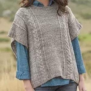 May include: A beige cable-knit poncho with short sleeves. The poncho features a textured pattern and is worn over a blue plaid shirt. The garment is made of a soft, warm material, perfect for layering during cooler weather.