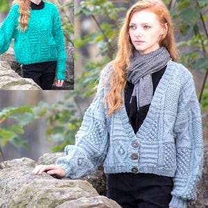 May include: A light blue cable-knit cardigan with a V-neck and three brown buttons. The sweater features intricate patterns and is worn with a black top and scarf. A teal cable-knit sweater is shown in the inset.