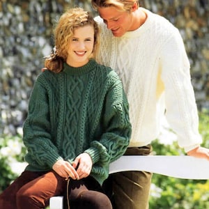 May include: A couple wearing matching cable knit sweaters. The woman is wearing a green sweater and the man is wearing a white sweater. They are standing in front of a stone wall.