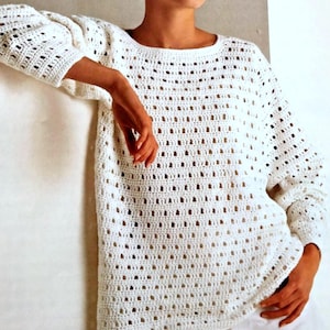 Könnte beinhalten: Ein weißer Häkelpullover mit offenem Gewebe und langen Ärmeln. Der Pullover hat einen runden Ausschnitt und eine lockere, bequeme Passform. Das Häkelmuster erzeugt ein strukturiertes, luftiges Design, perfekt zum Überziehen oder für wärmeres Wetter.