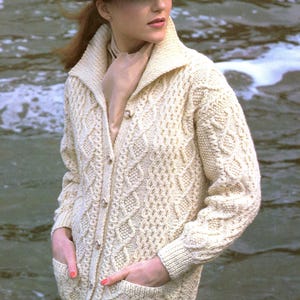 Op de afbeelding: Een witte kabelgebreide cardigan met een knoopsluiting en zakken. De cardigan heeft een brede kraag en wordt gedragen over een geruite rok.