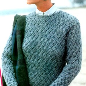 May include: A grey crew neck sweater with a basket weave pattern. The sweater has a textured appearance. A green and blue scarf is draped over the shoulder. The sweater is made of a soft material.