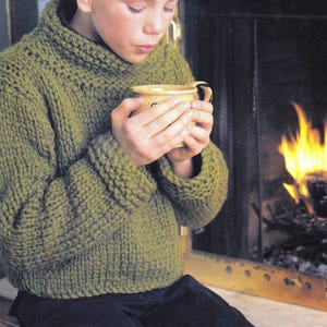 May include: A young person wearing a green knitted sweater sits in front of a fireplace, holding a yellow mug and looking down at it.