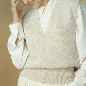 Puede incluir: Chaleco de punto color crema con cuello en V y cierre de botones. El chaleco tiene un patrón texturizado y se usa sobre una camisa blanca con cuello. La parte inferior del chaleco tiene un diseño acanalado.