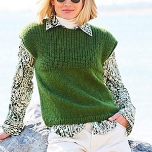 May include: A green knitted vest with a ribbed upper section and a smooth lower section. The vest is worn over a patterned long-sleeved shirt with a white collar. The outfit is completed with white trousers. The overall style is casual and comfortable.