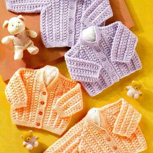 May include: A selection of hand-knitted baby cardigans in pastel shades. The image shows two lavender and two peach-coloured sweaters, each with button fastenings. Small teddy bear and piglet soft toys are placed around the sweaters.