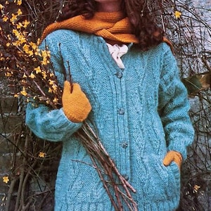 May include: A light blue cable-knit cardigan with a button-up front and pockets. The person is wearing a mustard-colored scarf and gloves, holding a bundle of branches with yellow flowers. The cardigan is a cozy, textured garment.