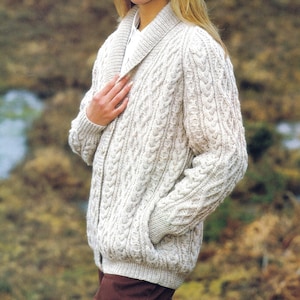 May include: A cream-colored cable-knit cardigan with a shawl collar and two front pockets. The sweater has a textured pattern and a button closure. The sleeves are long, and the overall style is classic and cozy.