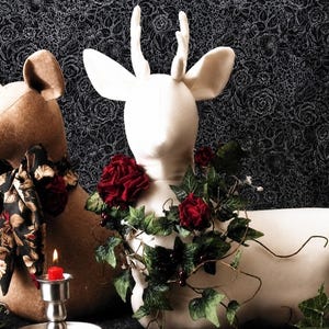 May include: Two fabric deer figures, one tan and one cream, featuring antlers. The tan deer wears a black floral patterned scarf. The cream deer is adorned with a garland of dark red roses, green ivy leaves, and small berries. A red candle sits in a silver holder.