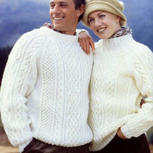 Puede incluir: Una pareja con suéteres de punto trenzado color crema a juego. Los suéteres tienen patrones intrincados y un diseño clásico y texturizado. El hombre lleva una camisa con cuello debajo. La mujer lleva un sombrero y una bufanda.