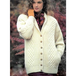 May include: Cream-colored cable-knit cardigan sweater with a button-down front and two front pockets. The sweater features intricate cable patterns and wooden buttons. The model is wearing a patterned scarf and orange gloves.