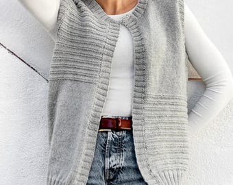 May include: A light gray knitted vest with a ribbed design. The vest is worn over a white long-sleeved shirt and blue jeans. A brown belt is visible. The vest has a round neckline and a sleeveless design.