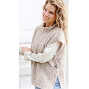 Op de afbeelding: Een beige, getextureerde gebreide vest met een hoge halslijn en zijknopen. Het vest is gelaagd over een wit shirt met lange mouwen. De algehele stijl is casual en comfortabel.