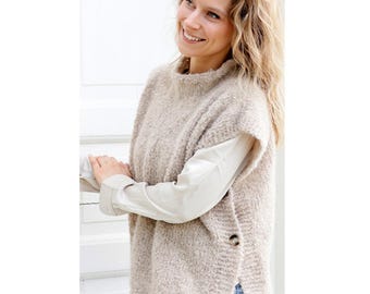 May include: A beige, textured knit vest with a high neckline and side buttons. The vest is layered over a white long-sleeved shirt. The overall style is casual and comfortable.