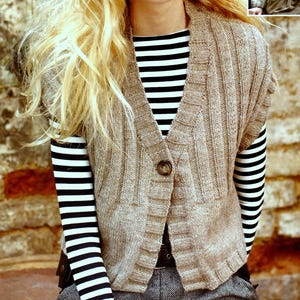 May include: A tan knitted vest with a single button closure. The vest has a ribbed texture and is worn over a black and white striped long-sleeved shirt. The background is a brick wall.