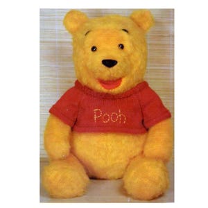 May include: A plush teddy bear with golden yellow fur, featuring a round head, small ears, and a black nose. The bear is wearing a short-sleeved red knitted sweater with the word Pooh embroidered in yellow thread across the center of the chest.