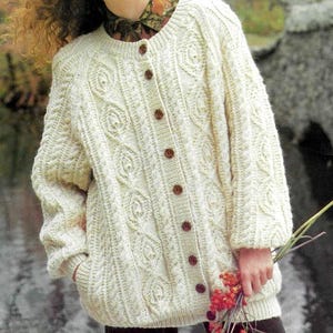 May include: Cream-colored, button-down cardigan sweater with a cable knit design. The sweater features a round neckline, long sleeves, and brown buttons. The cable knit pattern includes a rose-like motif. The sweater has pockets and is made of a thick, textured yarn.