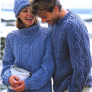Puede incluir: Una pareja con suéteres azules de punto trenzado a juego y un gorro. Los suéteres tienen un patrón texturizado y un cuello alto. La mujer sostiene un cubo blanco. El fondo sugiere un entorno de playa.