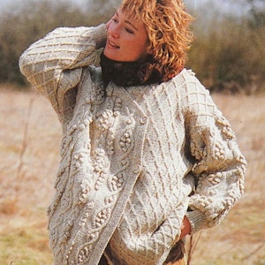 May include: A light beige, hand-knitted cardigan with intricate cable and bobble stitch detailing. The sweater features a wrap-style front with a single button closure and a textured pattern. The sleeves and body have a diamond-like pattern. The cardigan is a cozy, handmade garment.