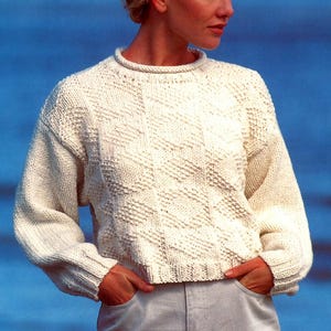 May include: Cream-colored knit sweater with a textured diamond pattern. The sweater has a crew neck and long sleeves with ribbed cuffs. The model is wearing the sweater with light-colored jeans. The background is a solid blue.