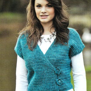 May include: Teal crochet wrap-style cardigan with short sleeves and flower-shaped buttons. The cardigan is worn over a white long-sleeved top. The crochet stitch creates a textured look. The wrap design adds a touch of elegance to the garment.
