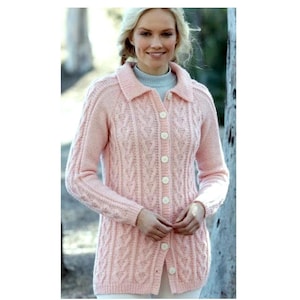 Könnte beinhalten: Hellrosa Cardigan mit langen Ärmeln, Kragen und weißen Knöpfen. Der Pullover hat ein Zopfmuster und wird über einem hellblauen Rollkragenpullover getragen. Die Frau lächelt.