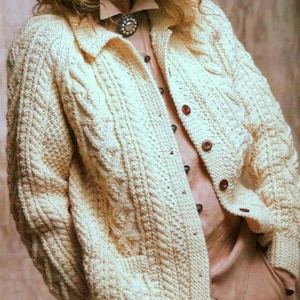 May include: Cream cable-knit cardigan with a collared neckline and wooden buttons. The textured knitwear features a classic cable pattern and a pocket on the left side. The cardigan is worn over a light brown blouse.