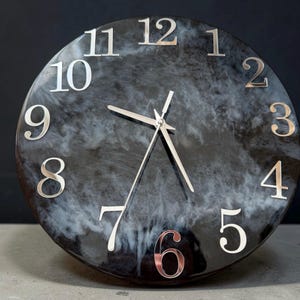 Könnte beinhalten: Eine runde Wanduhr mit einem glänzenden, schwarz-weiß marmorierten Harzzifferblatt. Die Uhr hat große, metallisch silberne Ziffern von eins bis zwölf am Rand und drei dünne, passende silberne Zeiger in der Mitte des Zifferblatts.
