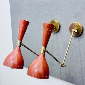 Op de afbeelding: Een paar rode en messing wandlampen met verstelbare armen. De lampen hebben een mid-century modern design en zijn voorzien van een wit gestippeld patroon.
