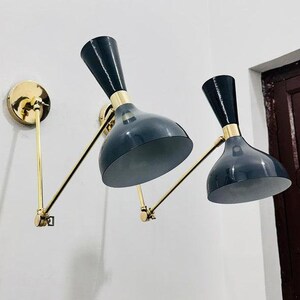 Op de afbeelding: Twee zwarte en gouden wandlampen met verstelbare armen. De lampen hebben een mid-century modern design met een zwarte kap en een gouden voet.