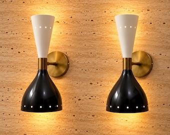 May include: A pair of wall sconces featuring a dual-cone design with a matte white upper cone and a glossy black lower dome. Each lamp has a brass-toned circular wall mount and a central brass connector, with small circular perforations.