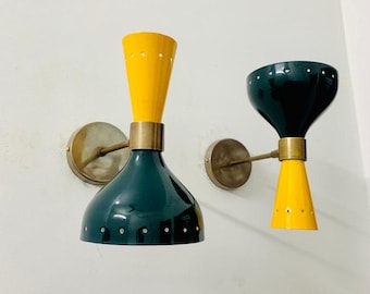May include: Two wall sconces with brass accents. The sconces have a yellow and dark green colour scheme. The shades are shaped like a cone and have a pattern of small holes.